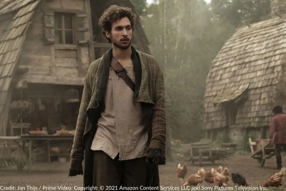 Barney Harris, as Mat Cauthon, walks through the Two Rivers, where chickens are feeding in the background and thatched-roof houses surround him.