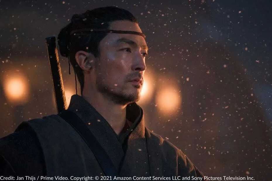 Daniel Henney as Lan Mandragoran stands resolutely amidst falling ash, his expression calm and focused, with a sword strapped to his back.