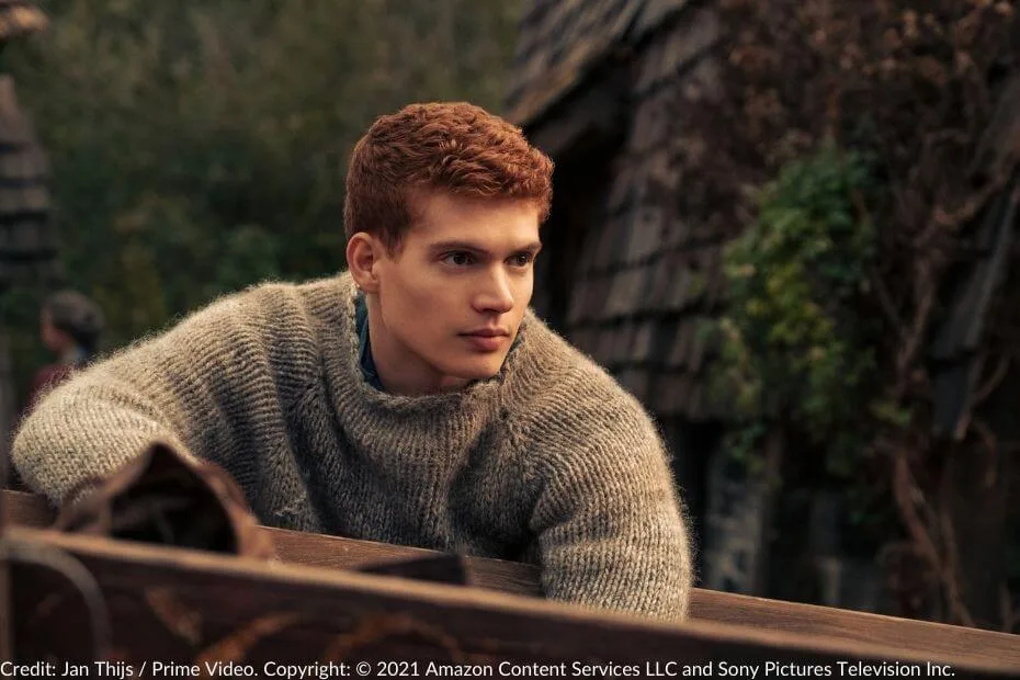 Josha Stradowski, as Rand al’Thor, dressed in a gray woolen jumper, leans thoughtfully against a wooden railing with rustic village homes in the background.