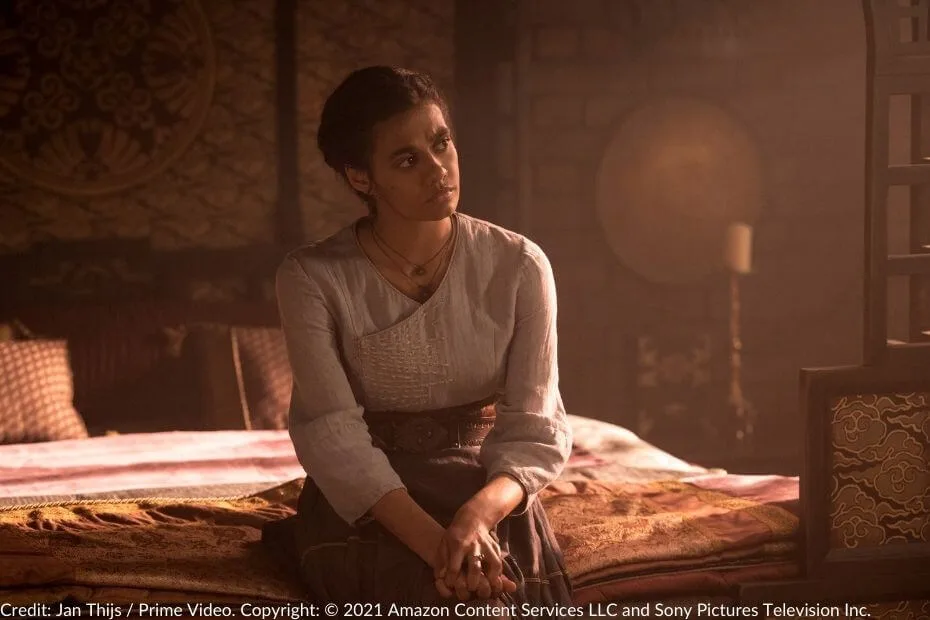 Madeleine Madden as Egwene al’Vere sits on the edge of a bed, her expression thoughtful, in a softly lit room with intricate decor.