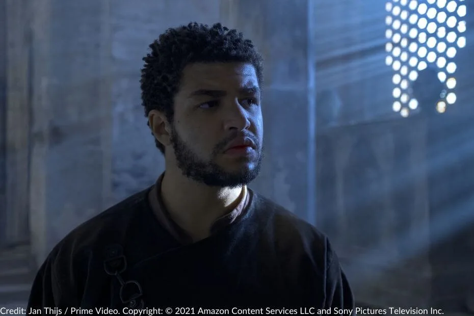 Marcus Rutherford, as Perrin Aybara, stands in a dimly lit room in Shadar Logoth, his expression tense and thoughtful as sunlight streams through a lattice window behind him.