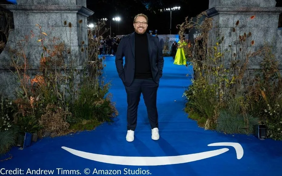 Rafe Judkins, Showrunner and Executive Producer of The Wheel of Time, stands on the vibrant blue carpet at the series’ London premiere, framed by floral arrangements and iconic set design elements inspired by the show.