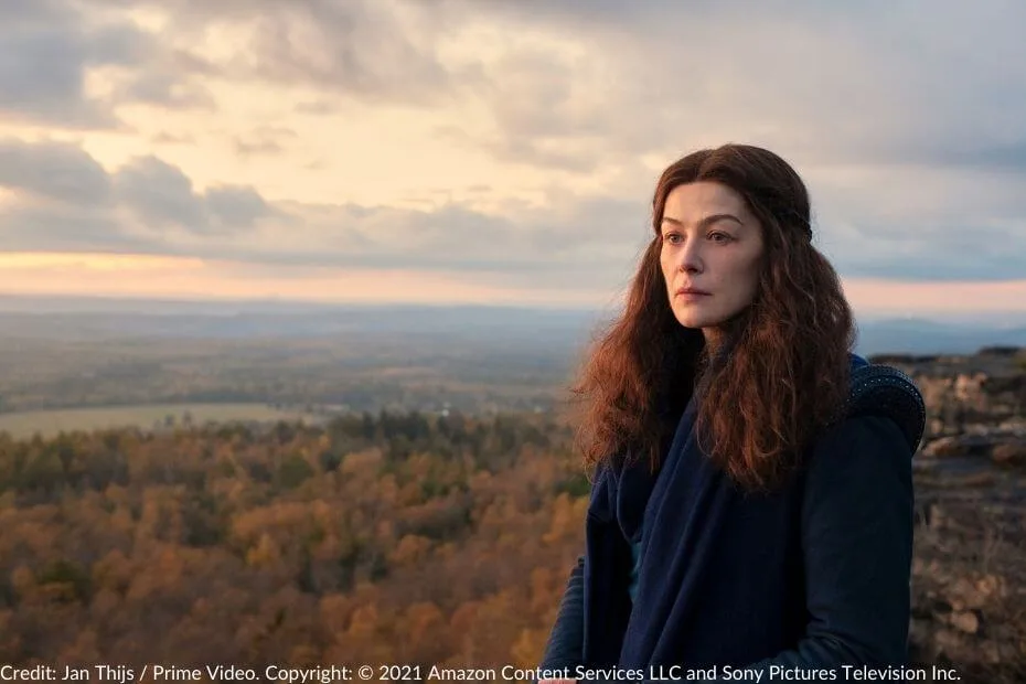 Rosamund Pike as Moiraine Damodred, standing solemnly on a cliff’s edge, overlooking a sprawling autumnal forest bathed in the warm hues of a setting sun.