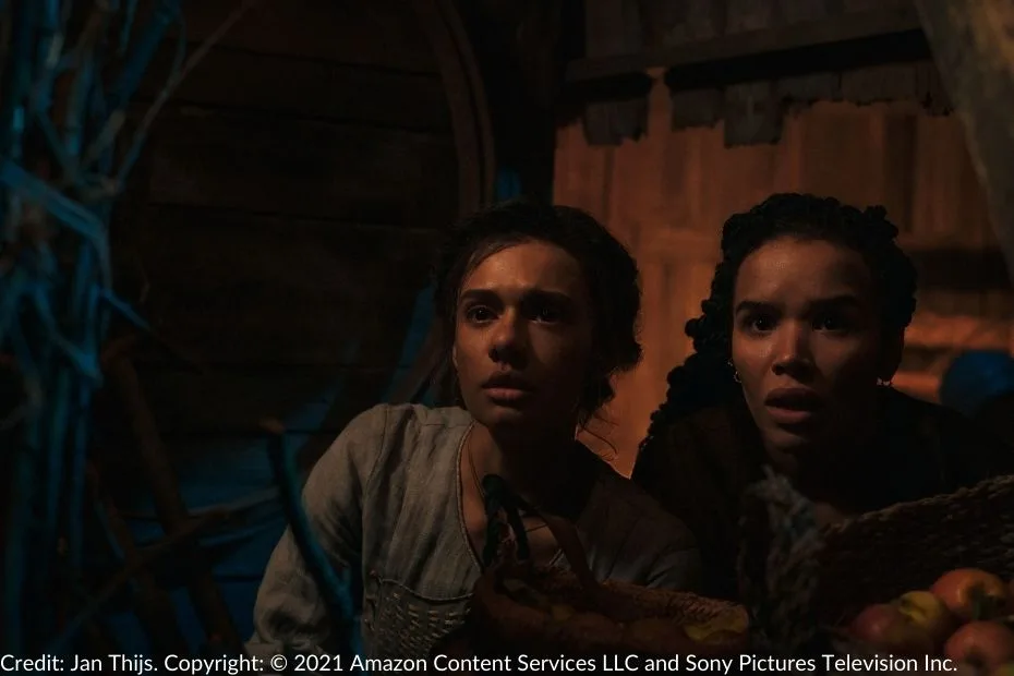 Egwene and Nynaeve crouch together in a dimly lit space, fear evident as they hide from the marauding Trollocs threatening their village. A basket of fruit and a rustic wooden backdrop frame the scene.