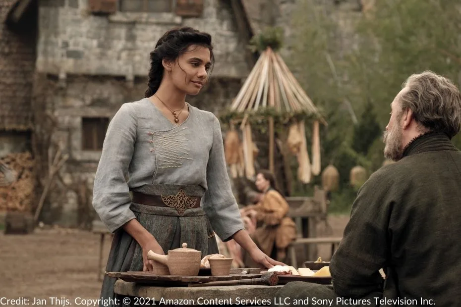 Egwene speaks with Tam at a wooden table in the village square. The table is set with clay pots and a plate of cheese.