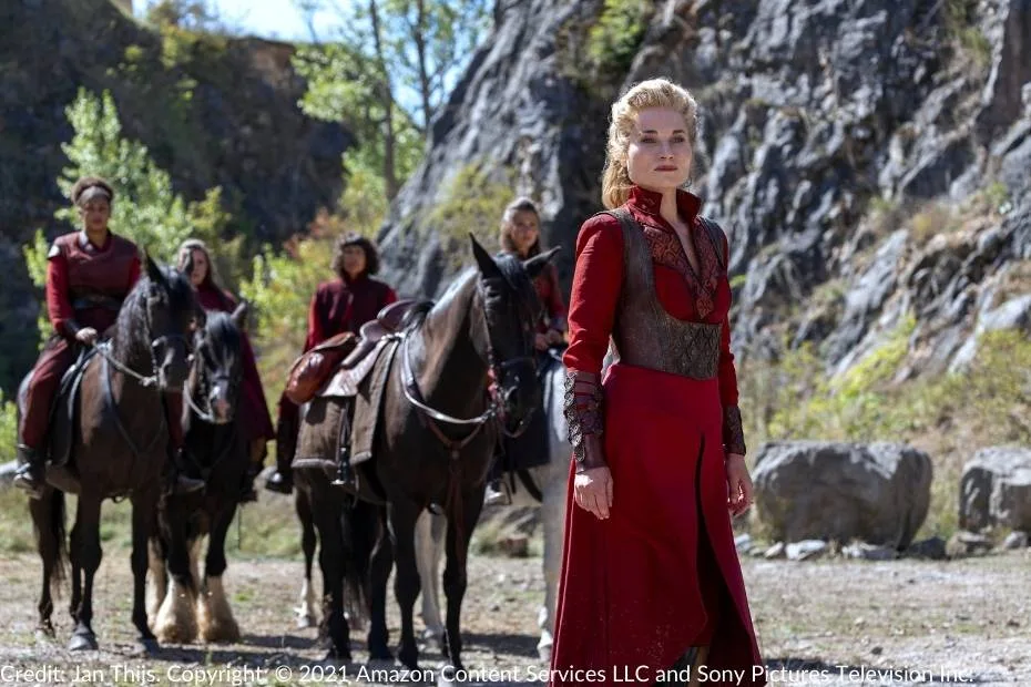 Liandrin Guirale stands confidently in her signature red attire, leading a group of mounted Red Sisters. Behind her, the other Aes Sedai are seated on horseback, their outfits matching Liandrin’s vibrant red. The scene is set against a rocky and sunlit wilderness backdrop.