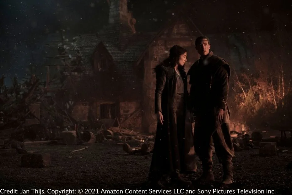 Moiraine stands wearily beside Lan amid the devastation of a crumbled building. The scene is dark, lit by the glowing embers of fires burning in the background.