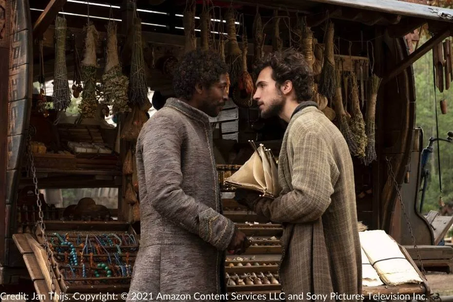 Mat Cauthon leans to speak with Padan Fain at the peddler’s wagon. Mat holds a pair of lanterns, while the wagon is adorned with colorful trinkets, herbs, and other goods for trade.