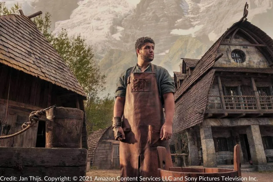 Perrin Aybara standing outside the village forge in the Two Rivers, wearing a leather apron and gloves. He looks off into the distance with a serious expression, framed by rustic buildings and the imposing mountains behind the village.