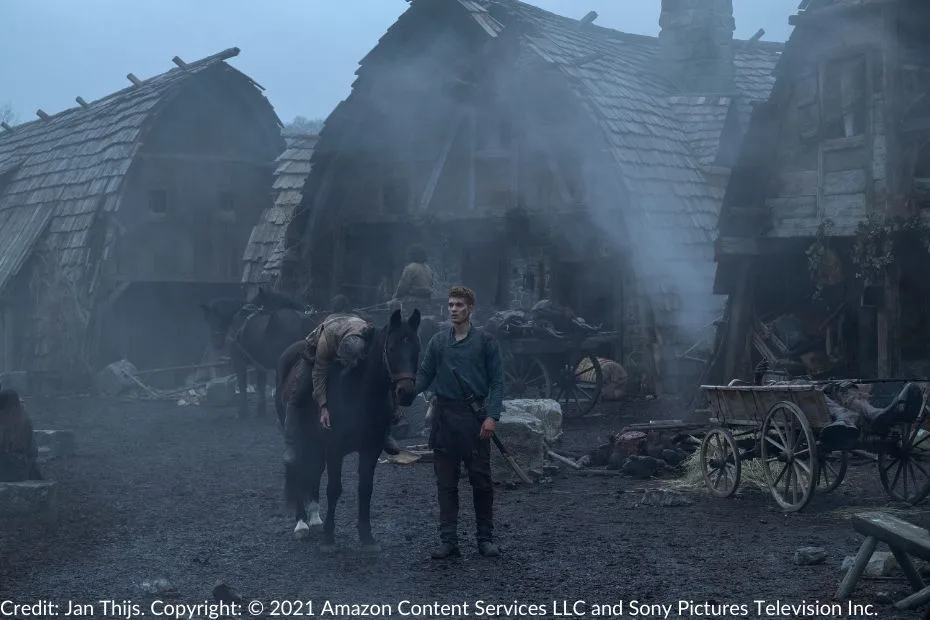 Rand walks alongside Bela, the mare, carrying a wounded Tam al’Thor, draped over the horse’s back. The scene is set in the aftermath of a Trolloc attack, with smoke filling the air and the village in ruins around them.