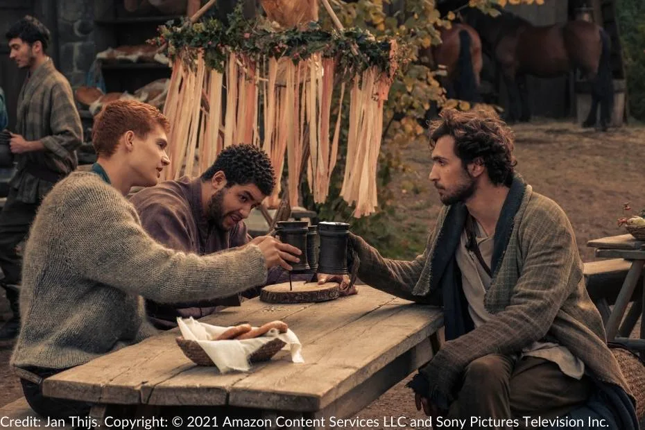 Rand, Perrin, and Mat sit at a wooden table outside, raising mugs of ale. Festive decorations and horses are in the background.