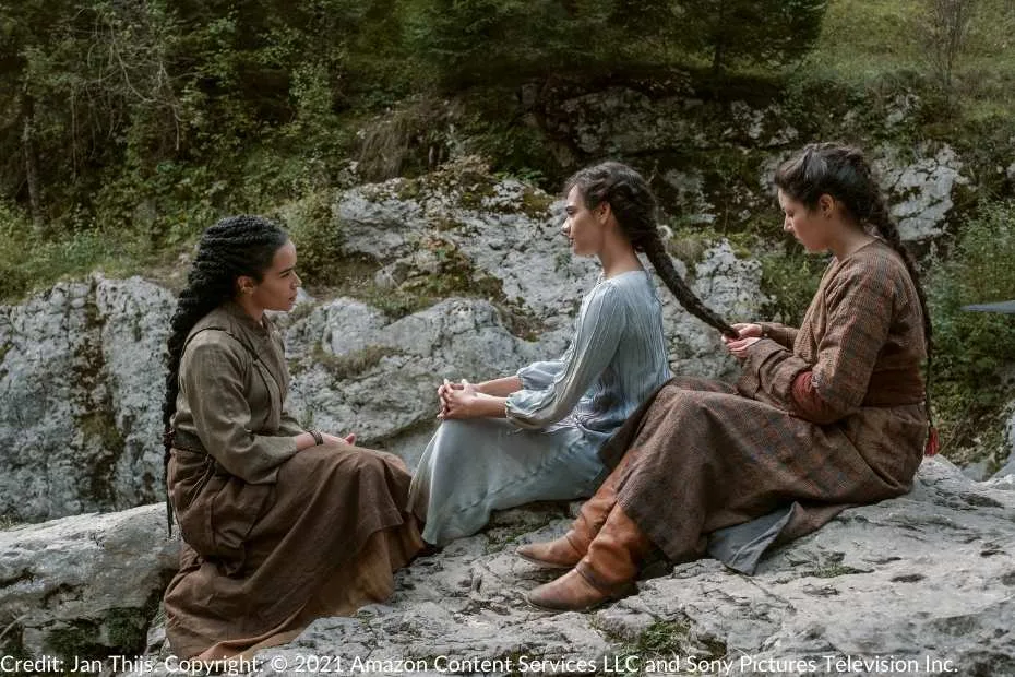 Egwene sits on a rocky clifftop while her hair is braided during the initiation ceremony. Nynaeve sits opposite Egwene, speaking to her calmly and reassuringly, while another woman works on Egwene’s braid. The serene mountain setting enhances the intimate moment.