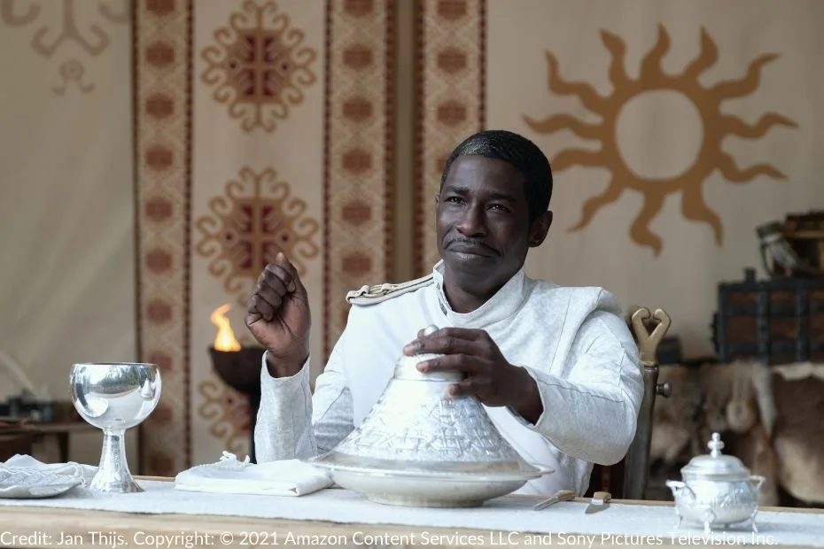 Eamon Valda sits at an elegantly set table dressed in the signature white of the Children of the Light. He holds the lid of a silver dish as if about to reveal its contents. A silver goblet, delicate white napkin, and intricate tableware add to the refined yet unsettling atmosphere. Behind him, the tent displays a golden sunburst, an emblem of the Whitecloaks.