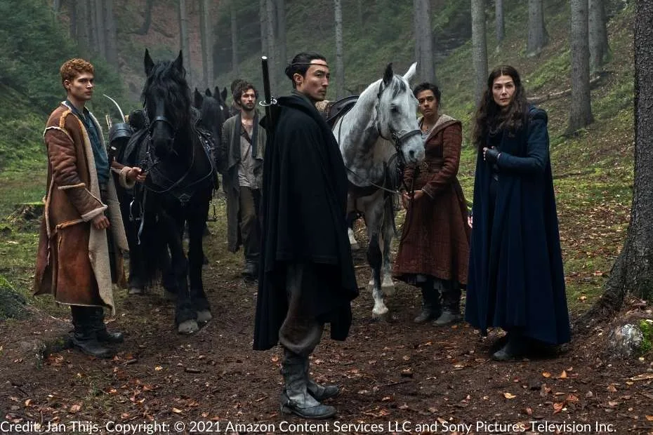 Rand, Mat, Lan, Egwene, and Moiraine stand together in a forest clearing, their horses nearby. The characters are dismounted and tense as if preparing for an imminent encounter.