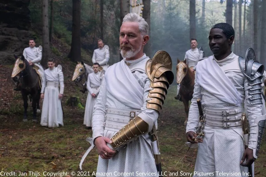 Geofram Bornhald and Eamon Valda stand at the forefront of a group of Whitecloaks in a forest clearing. Bornhald is clad in white robes with golden armor, while Valda’s expression is calm but commanding. Mounted Whitecloak soldiers and others standing in formation are visible in the background.