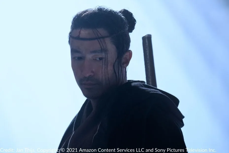 Daniel Henney wears a traditional leather hadori headband and gazes downward solemnly. His sword is visible on his back, and soft light surrounds him.