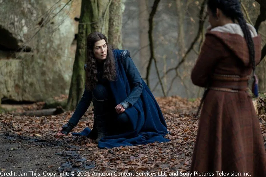 Moiraine kneels on the forest floor, one hand outstretched as if sensing the earth for something unseen. Egwene stands in the foreground, curiously observing Moiraine’s actions..