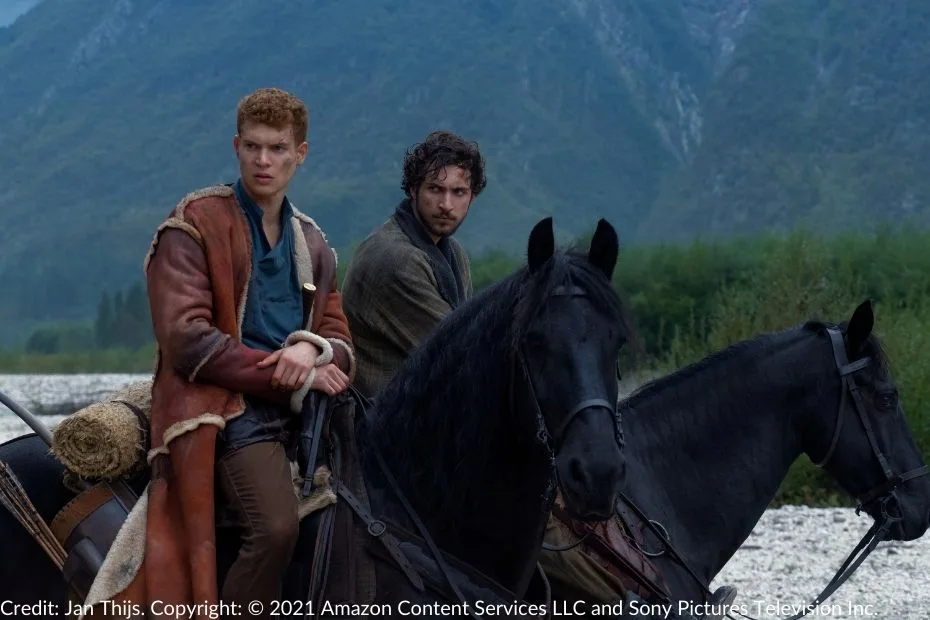 Rand and Mat sit atop their horses, gazing warily into the distance. The scene is framed by a riverbank and lush greenery, with mountains rising in the background.