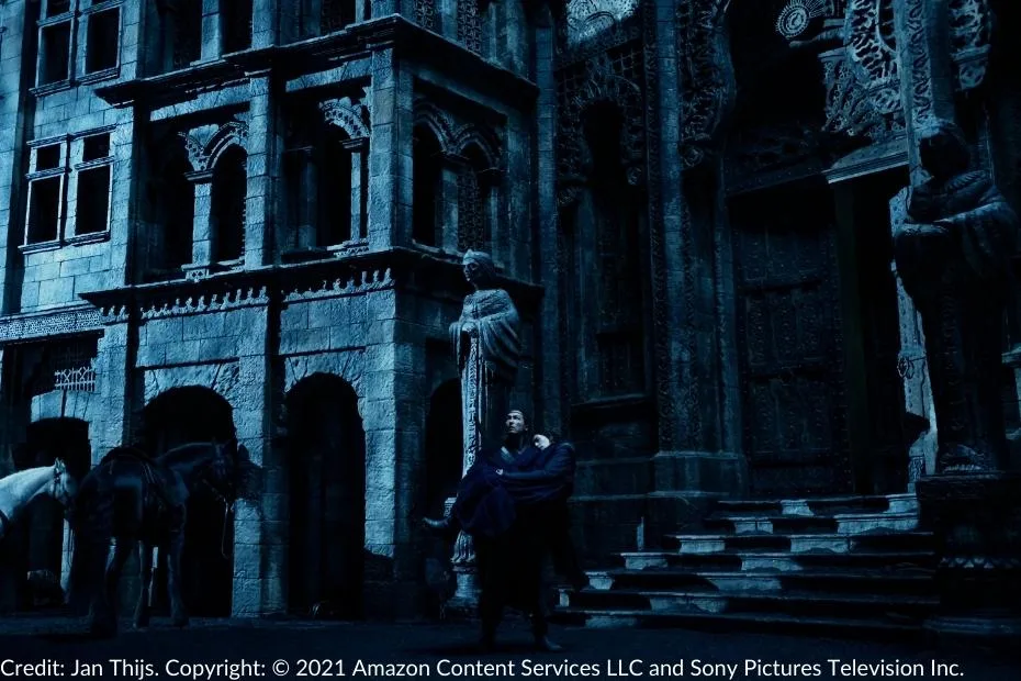 Lan carries an unconscious Moiraine in his arms as he exits a shadowy, ominous building in Shadar Logoth. The dark blue tones highlight the eerie architecture adorned with intricate carvings, foreboding statues, and horses waiting in the shadows.