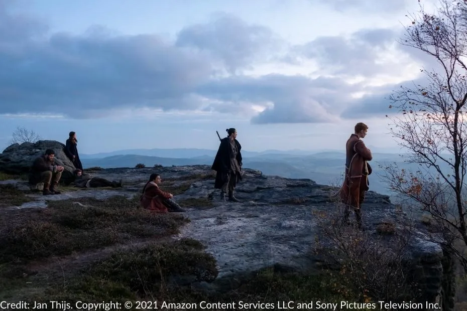 Lan, Moiraine, Rand, Mat, Perrin, and Egwene rest atop a rocky cliff at sunset, gazing over the expansive rolling hills and valleys.