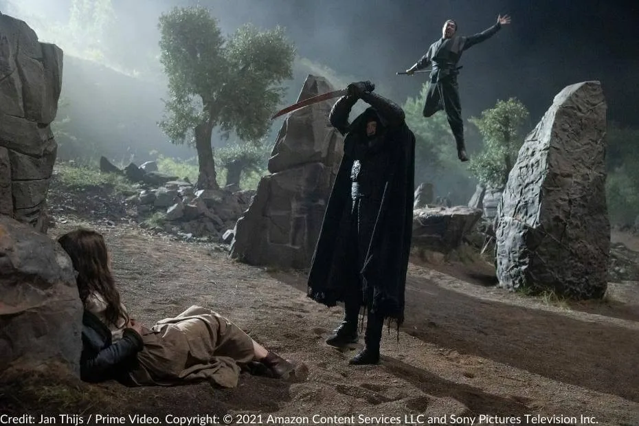 Behind-the-scenes image from The Wheel of Time Season 2, featuring Rosamund Pike as Moiraine Damodred on the ground, Daniel Henney as Lan Mandragoran leaping into action with his sword, and a Fade preparing to strike.