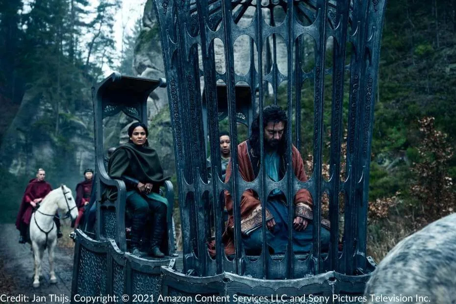 Alanna Mosvani sits on a wagon beside a caged Logain Ablar. The ornate metal cage confines Logain as Aes Sedai and their Warders escort him through a wooded trail.