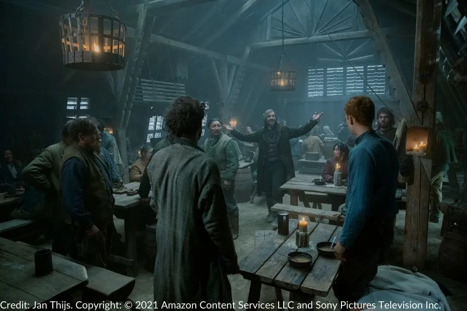 Thom Merrilin stands in the middle of the Four Kings Inn, his arms spread wide, smiling warmly. The inn is filled with patrons seated at wooden tables, illuminated by hanging lanterns and scattered candlelight.