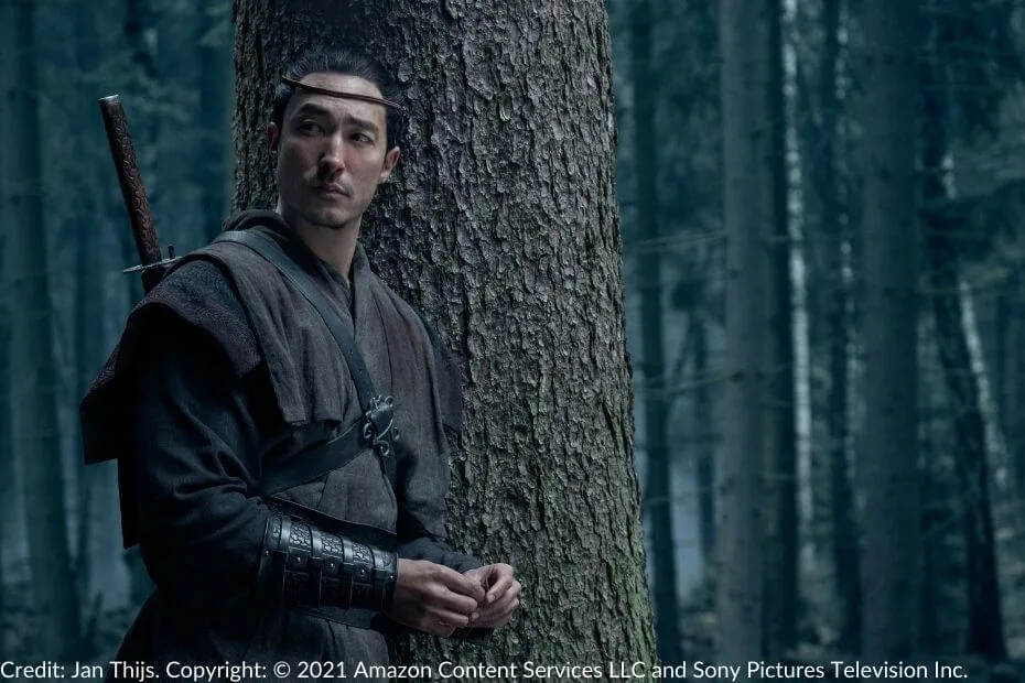 Lan Mandragoran leans against a tree in a dense forest. His sword is sheathed on his back, and he wears a leather headband (hadori) and sturdy travel attire. His expression is contemplative.