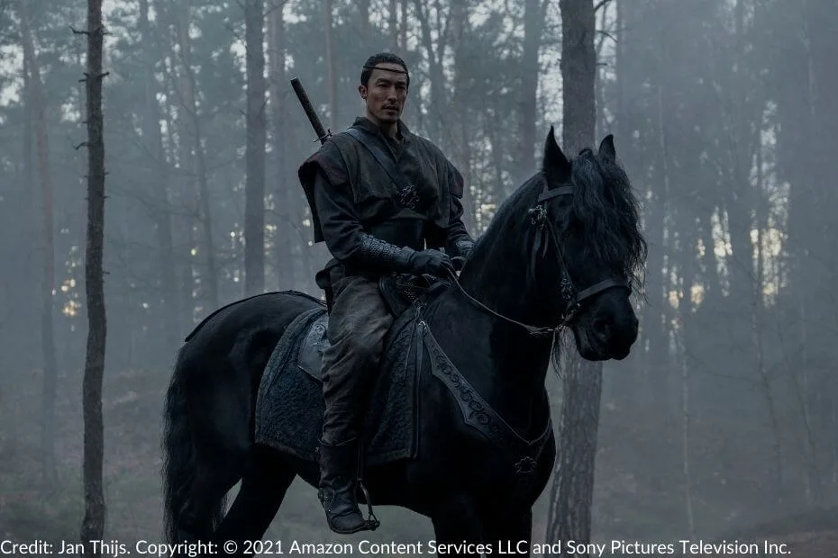 Lan sits atop a black horse in a misty forest. He wears dark, rugged travel attire, including a leather headband, with his sword on his back.