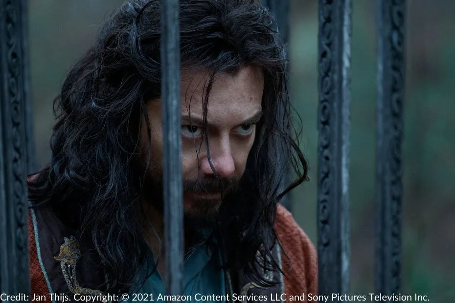 Logain peers intently through the bars of his cage. His disheveled hair and intense gaze convey defiance and determination.