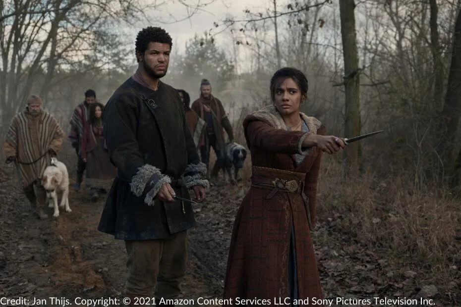 Perrin stands tensely, holding a blade at his side, while Egwene points a knife ahead with a nervous expression. A group of Tinkers and their dogs are behind them on a forest path.