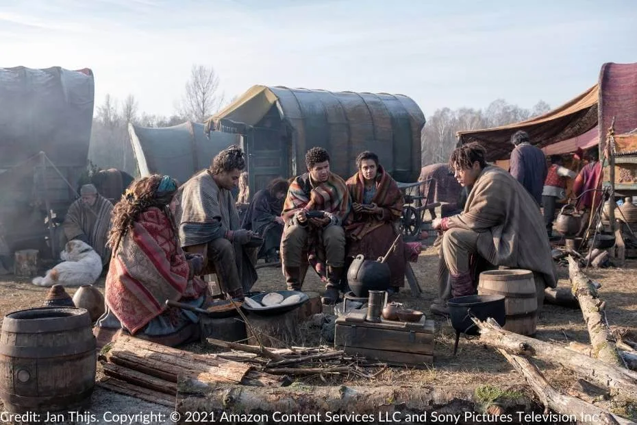 Perrin and Egwene sit wrapped in colorful blankets, eating and conversing with the Tinkers around a campfire. The camp has wagons and makeshift cooking areas in an open field under clear skies.