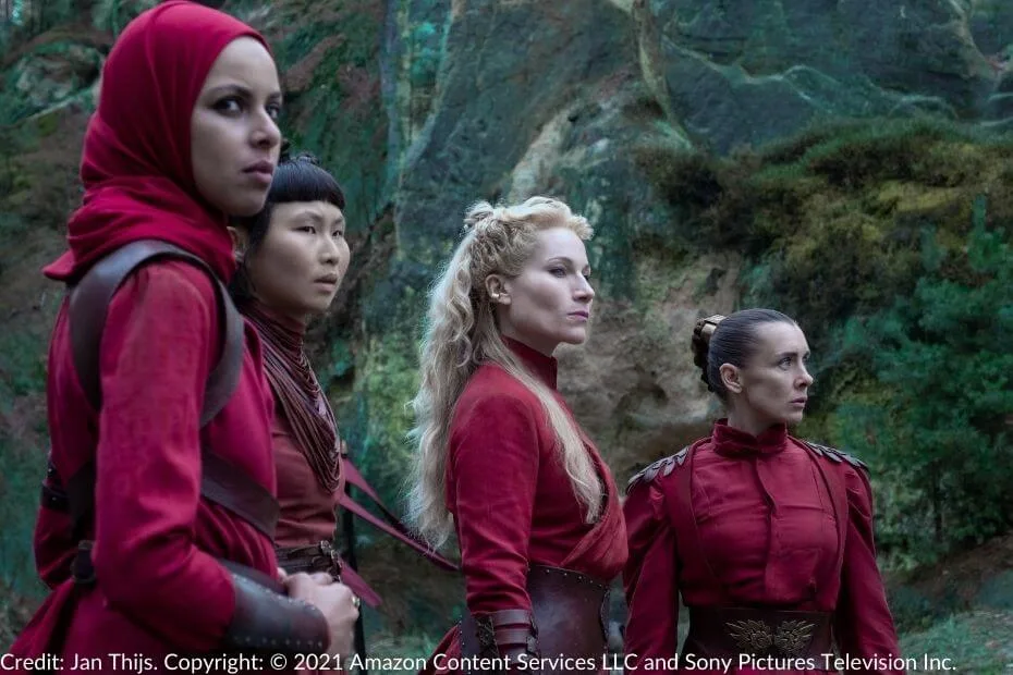 Liandrin leads three Red Sisters in a moment of gravity at the Aes Sedai camp. Dressed in deep crimson robes and leather accents, they stand against a rugged green and stone backdrop, their expressions reflecting determination and purpose.