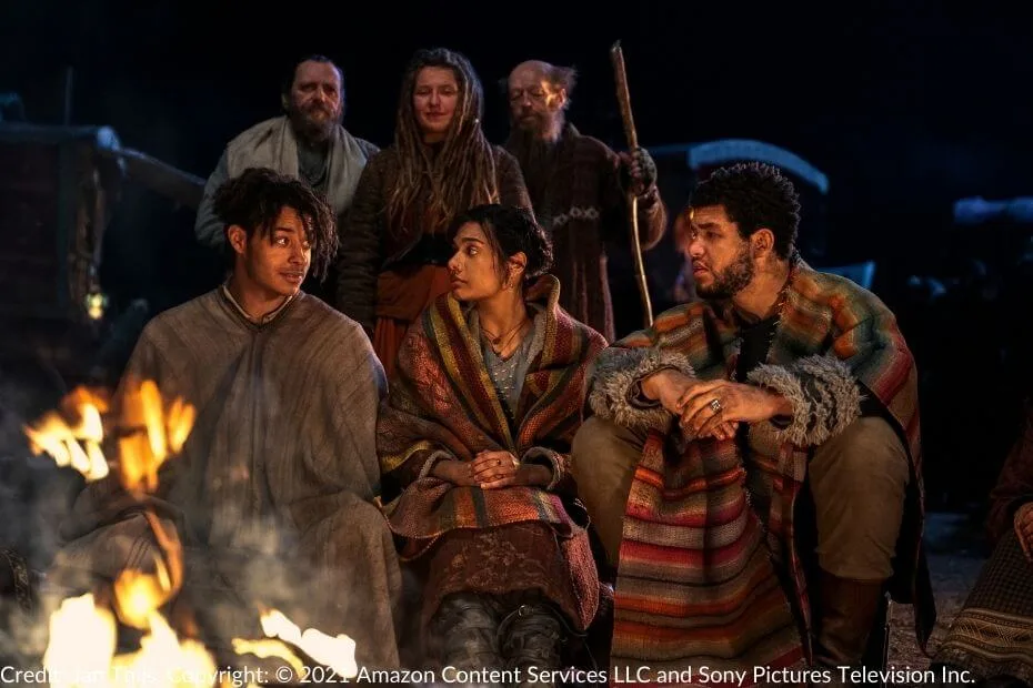 Aram, Egwene, and Perrin sit by a campfire wrapped in colorful blankets. Behind them, Tinkers stand and watch, illuminated by the warm firelight.