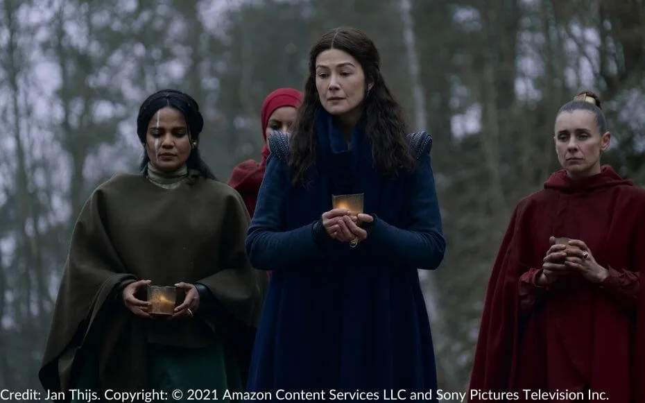 Alanna and Moiraine hold candles during a mourning ceremony. Two Red Sisters stand solemnly behind them, also holding candles, as the group honors the memory of Kerene Nagashi.