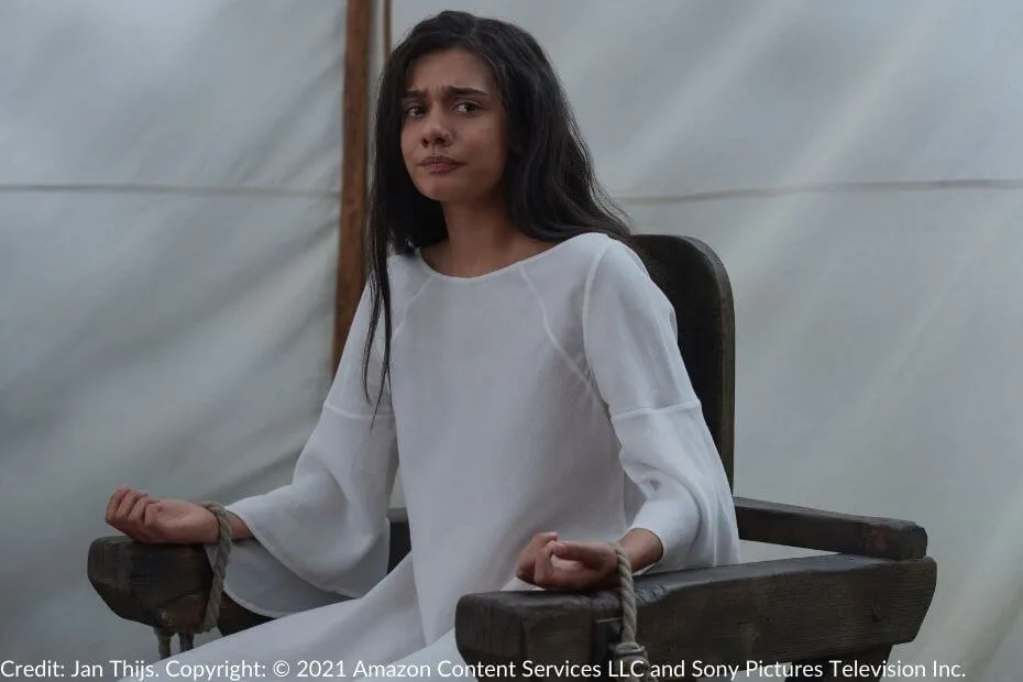 Egwene sits restrained, her wrists tied to a wooden chair. She wears a plain white gown and shows a mixture of fear and resolve.