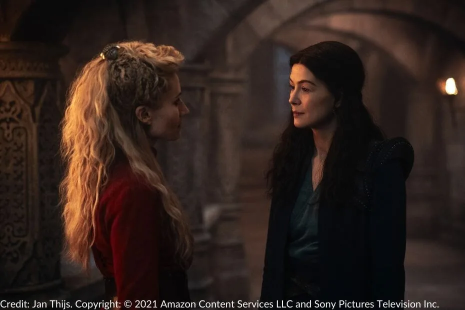 In a White Tower hallway, Liandrin, wearing a red dress, faces Moiraine, who wears blue.