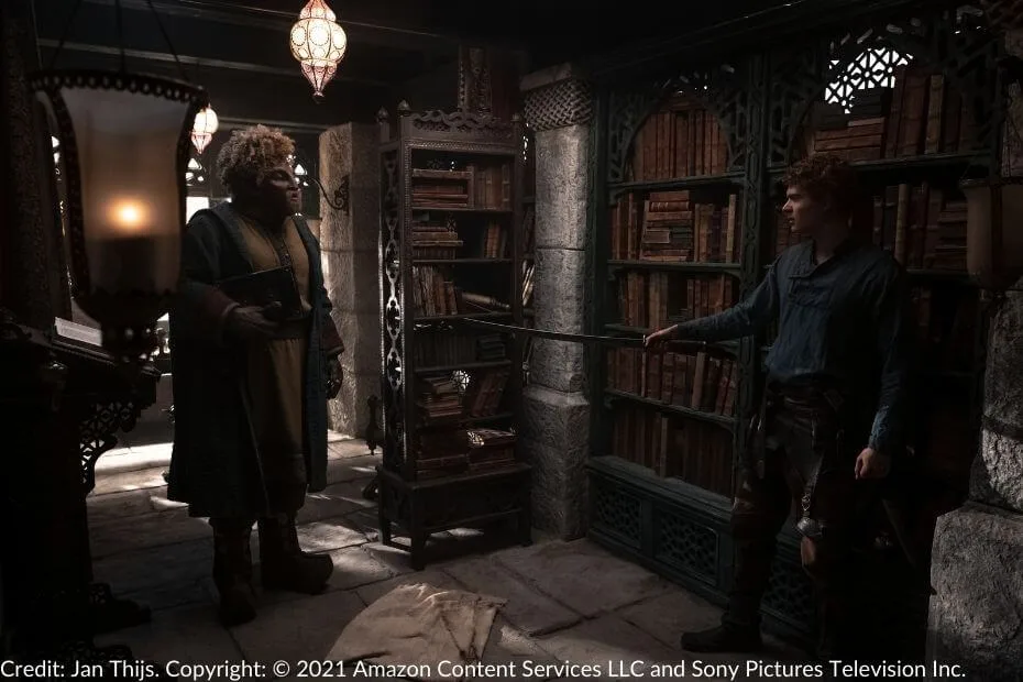 Rand cautiously holds his sword aloft as he encounters Loial in a dimly lit library filled with ancient books and ornate wooden shelves.