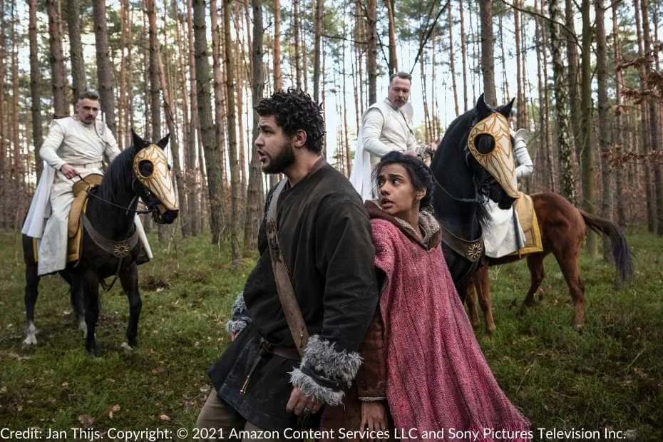 Perrin and Egwene stand back-to-back, tense and frightened, as two Whitecloaks on horseback loom behind them in a forest clearing.