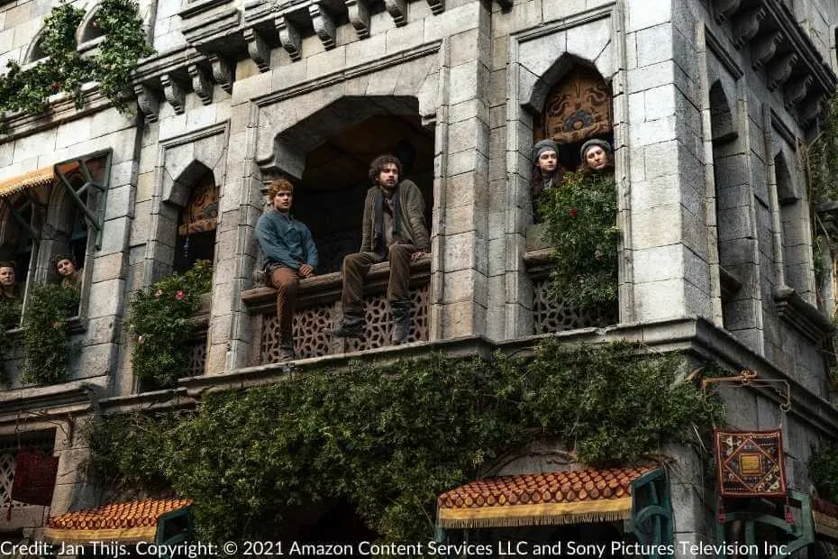 Rand and Mat sit on a balcony while other Tar Valon residents lean out of their windows in curiosity.