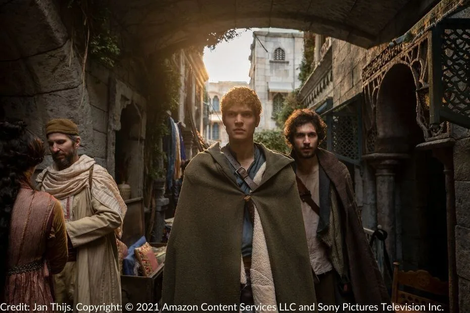 Rand and Mat, wearing cloaks, walk through a sunlit, narrow street in Tar Valon.