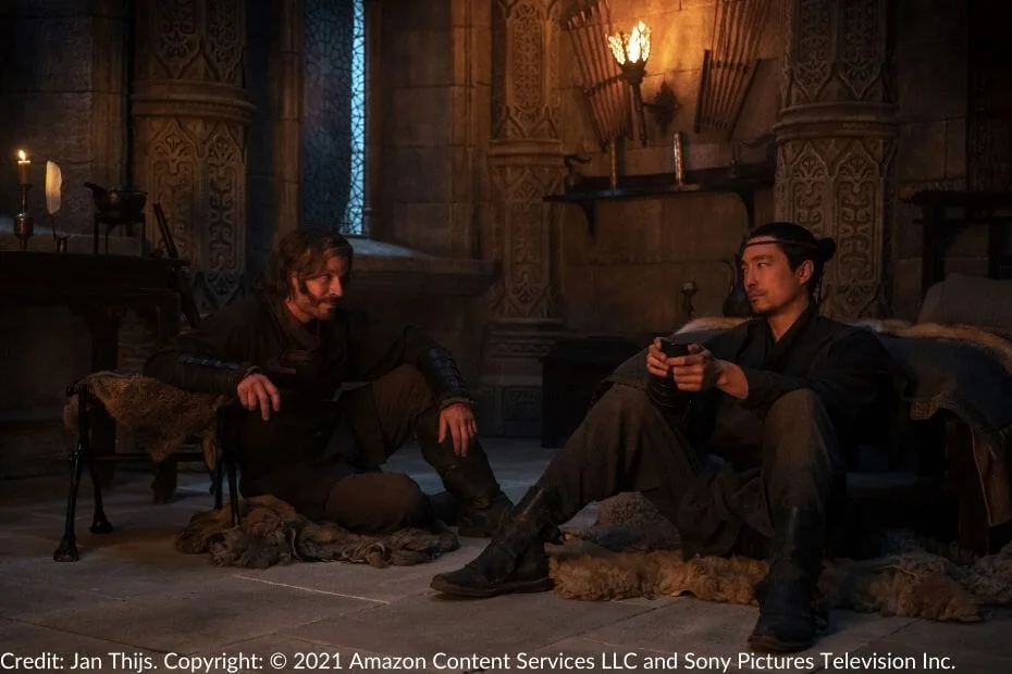 Stepin and Lan sit on rugs in a candlelit room, engaged in a somber conversation.