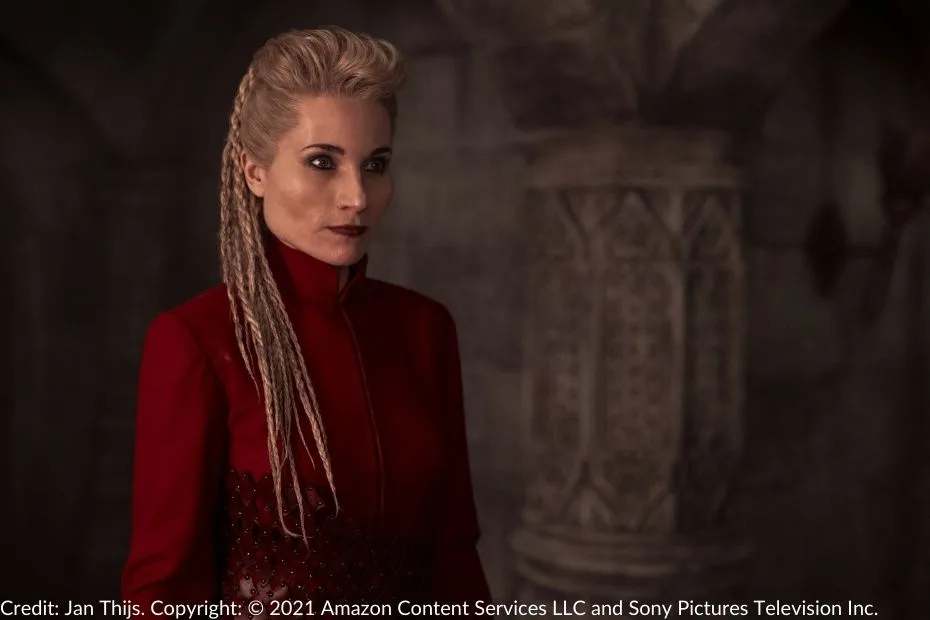 Liandrin stands in the White Tower, wearing a striking red dress with braided hair. Her expression is fierce and determined.