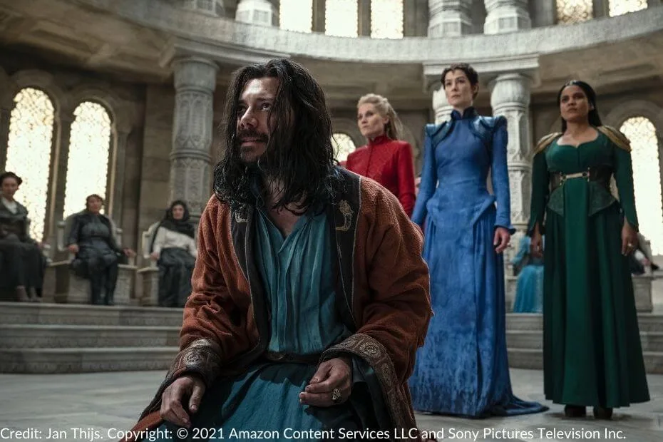 Logain kneels in the Hall of the White Tower, his expression defiant. Liandrin, Moiraine, and Alanna stand behind him, with other Aes Sedai seated in judgment on the raised perimeter.