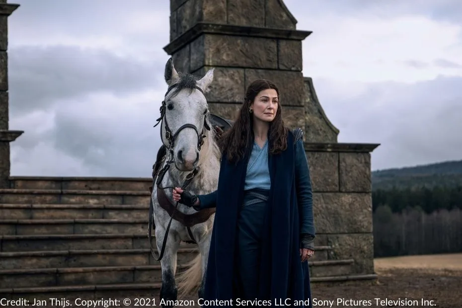 Moiraine stands beside her gray mare, Aldieb, outside the ancient stone structure of the Waygate, set against a cloudy sky and forested landscape.