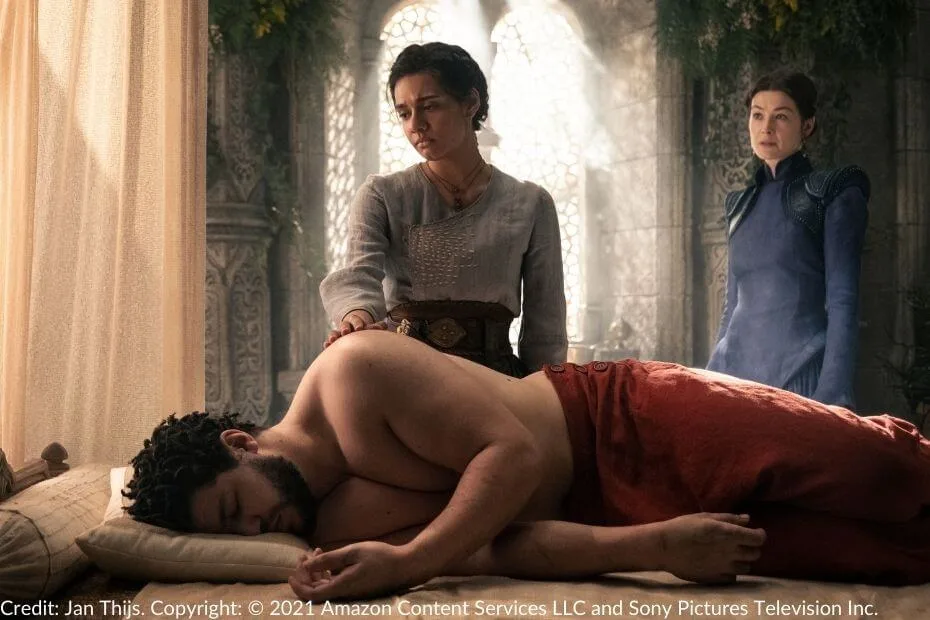 Egwene gently rests her hand on Perrin’s back, who lies shirtless on a bed. Moiraine observes from the background.