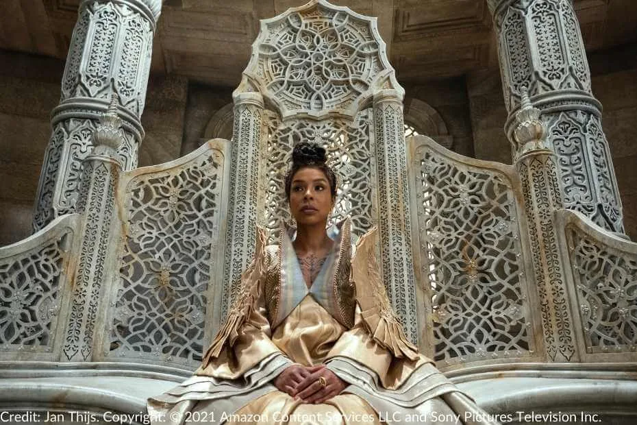 Siuan sits on the ornate Amyrlin Seat, dressed in gold ceremonial robes, with her hands resting calmly on her lap.