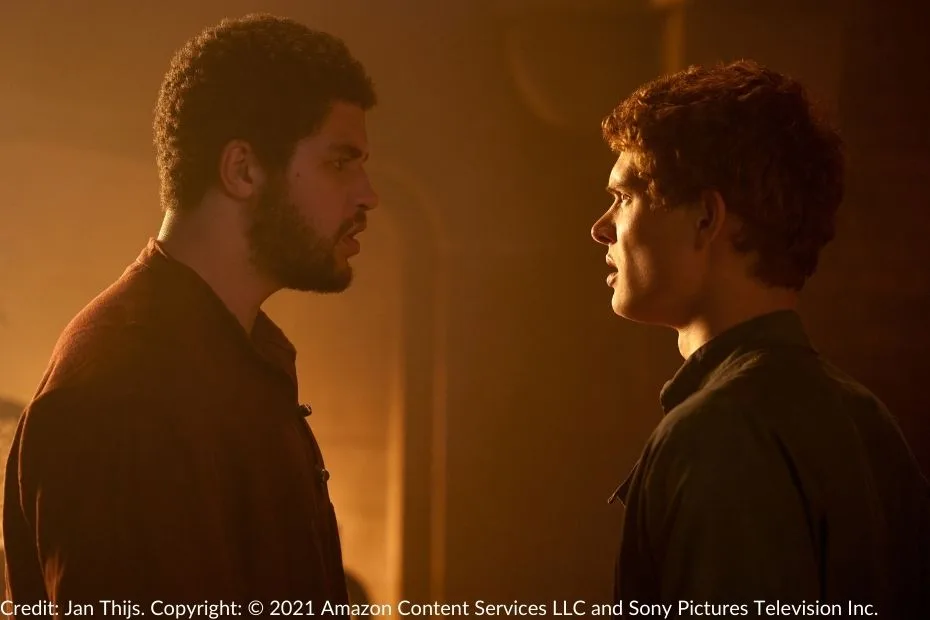 Perrin and Rand stand face-to-face in a dimly lit room, their expressions serious and conflicted. The warm light highlights their profiles as they confront one another.