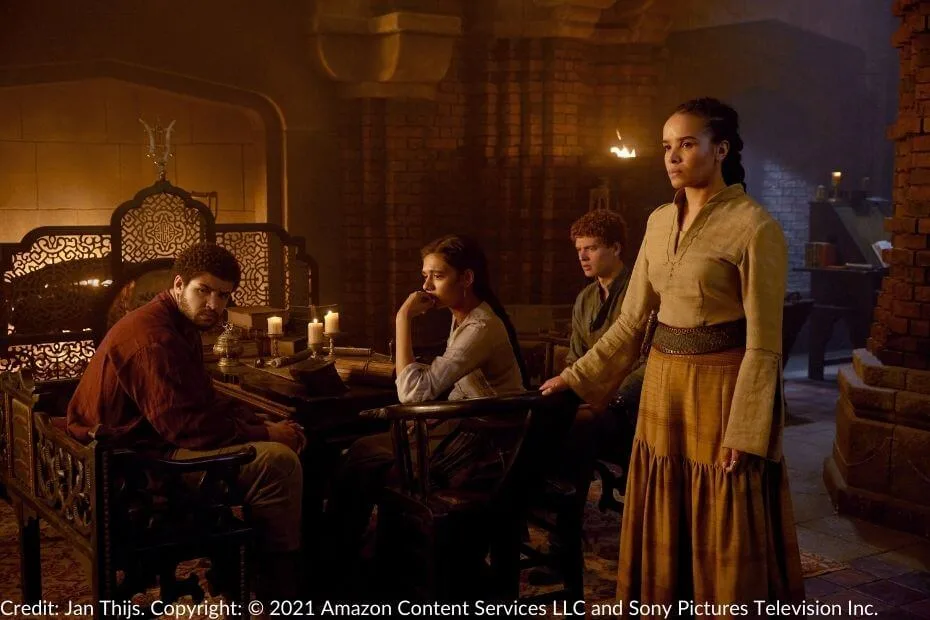 Perrin, Egwene, Rand, and Nynaeve in a dimly lit room. Nynaeve stands with a hand on her chair while the others sit silently around the table, their expressions heavy with concern.