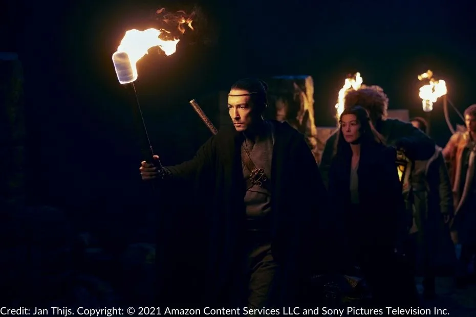 Lan holds a burning torch and cautiously leads the group through the dark and ominous Ways. The party follows behind him, their faces illuminated by the flickering firelight.